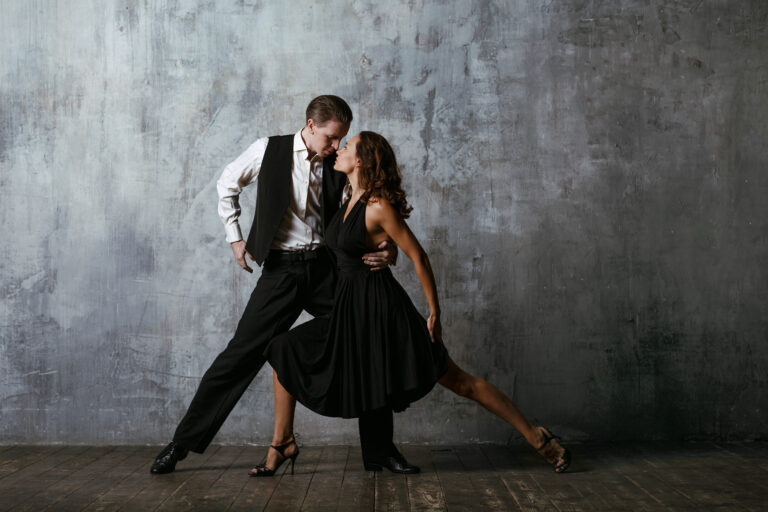 Couple dancing the tango in black-and-white outfits against a textured gray wall.