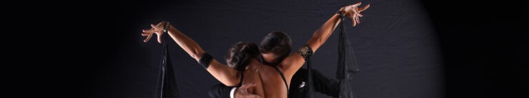 Couple in formal wear dancing the tango against a dark background, the woman lifting a flowing scarf.