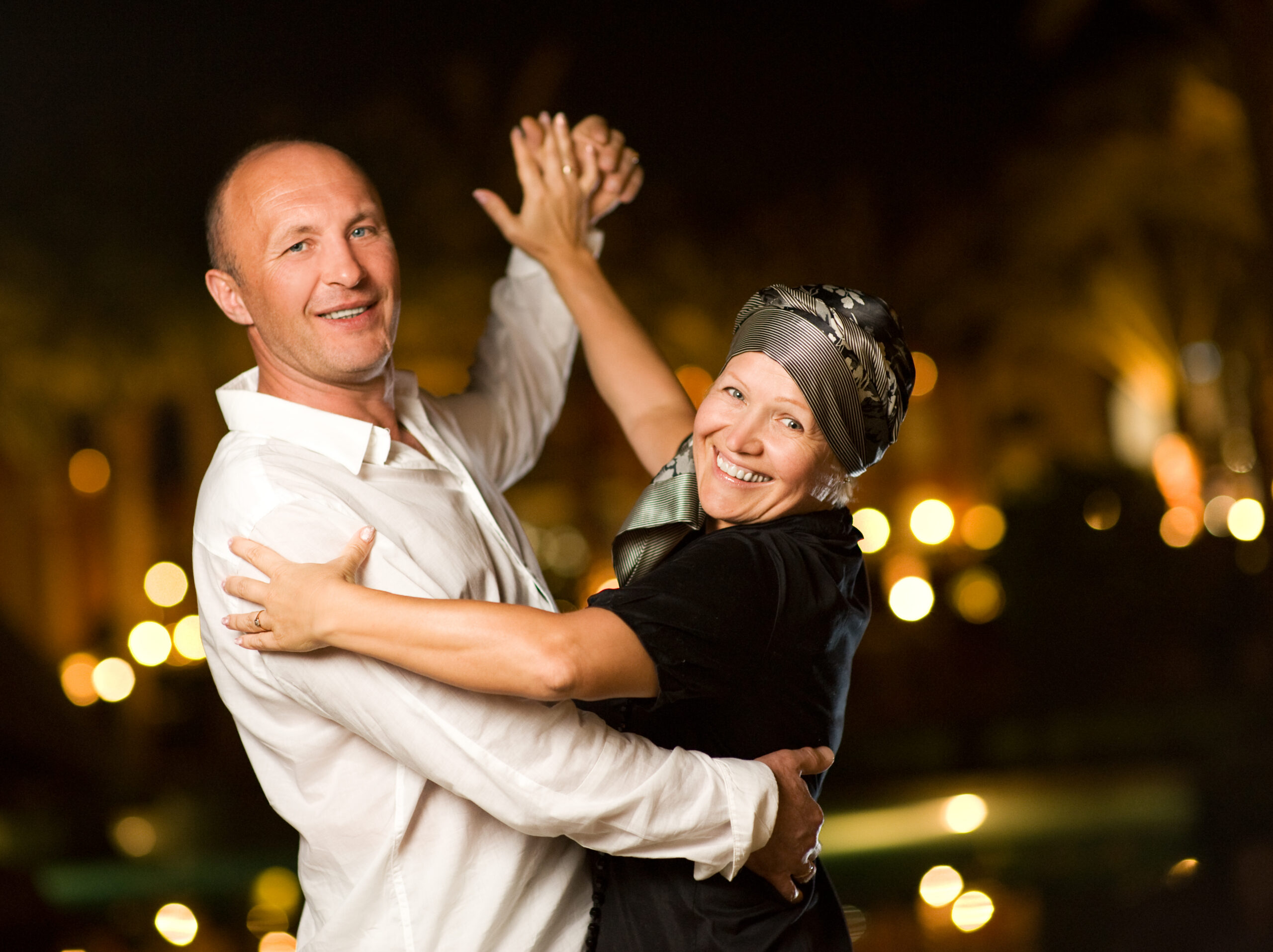 Couple dancing at night against blurred city lights, hands raised in a celebratory hold.