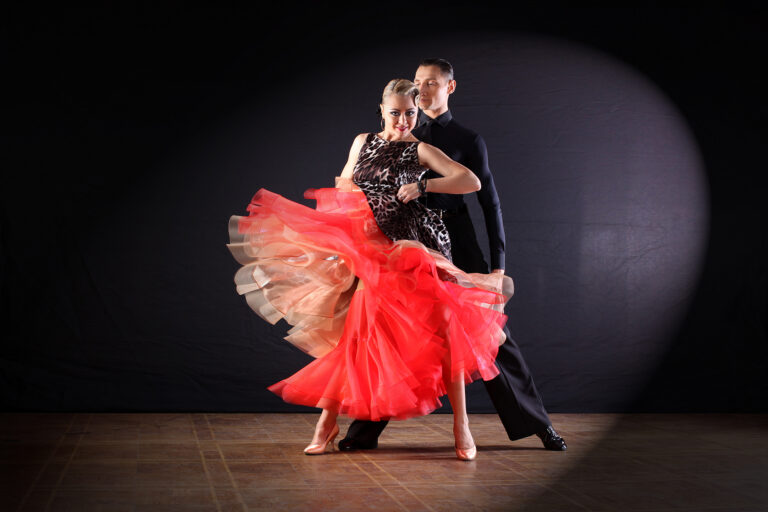Ballroom dance couple mid-turn on a dark stage, the woman in a leopard-print dress with a coral-red skirt.